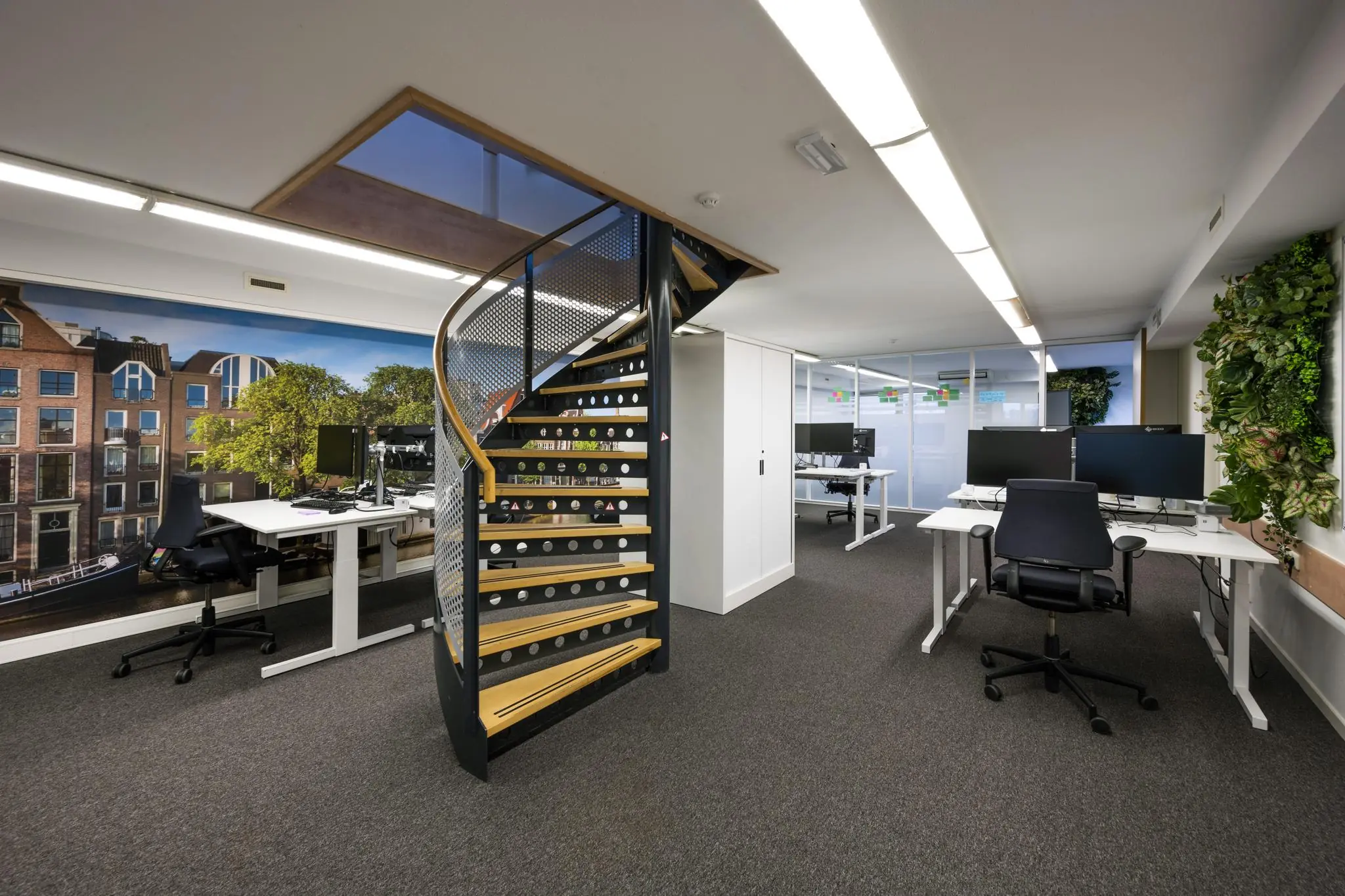 Modern kantoorinterieur aan de Prins Hendrikkade met werkplekken, een wenteltrap en een grote muurprint van Amsterdamse huizen.